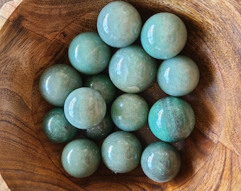 May include: A collection of polished, green aventurine spheres of varying sizes, displayed in a carved wooden bowl. The spheres have a smooth, reflective surface and range in shades from light to deep green.