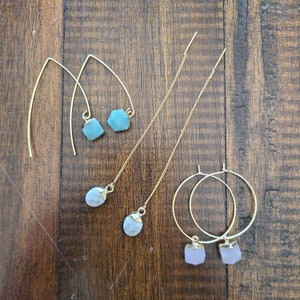 May include: An assortment of gold-toned earrings with gemstone accents. The collection includes drop earrings with blue-green, white, and lavender-colored stones, as well as hoop earrings and threader earrings. The earrings are displayed on a wooden surface.