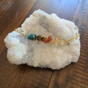 May include: A gold chain bracelet featuring a row of colorful, raw gemstones. The stones include shades of blue, green, brown, orange, and yellow. The bracelet is displayed on a white, crystalline surface, with a wooden background.