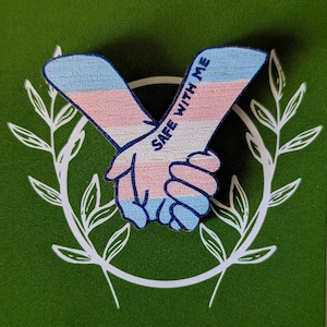 May include: A pin showing two hands clasped, coloured in the transgender flag's colours. The pin is on a green card with the text "SAFE WITH ME" and "Vicki Jordan Art". The website address is also visible.