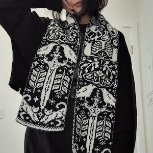 May include: A black and white knitted scarf with a repeating pattern of stylized figures and shapes. The scarf is draped over a black outfit. The design features a symmetrical arrangement of various elements, creating a visually striking accessory.