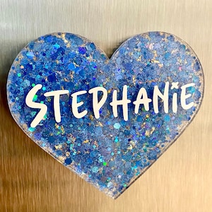 Puede incluir: Un adorno de resina azul en forma de corazón con purpurina dorada incrustada en la resina. El nombre "Stephanie" está escrito en letras blancas en el adorno.