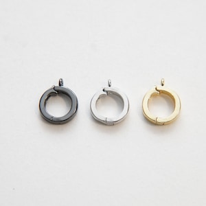Op de afbeelding: Drie metalen ringgespen in verschillende afwerkingen: zwart, zilver en goud. Elke gesp heeft een ronde vorm met een kleine opening om een ​​ketting of koord aan te bevestigen.