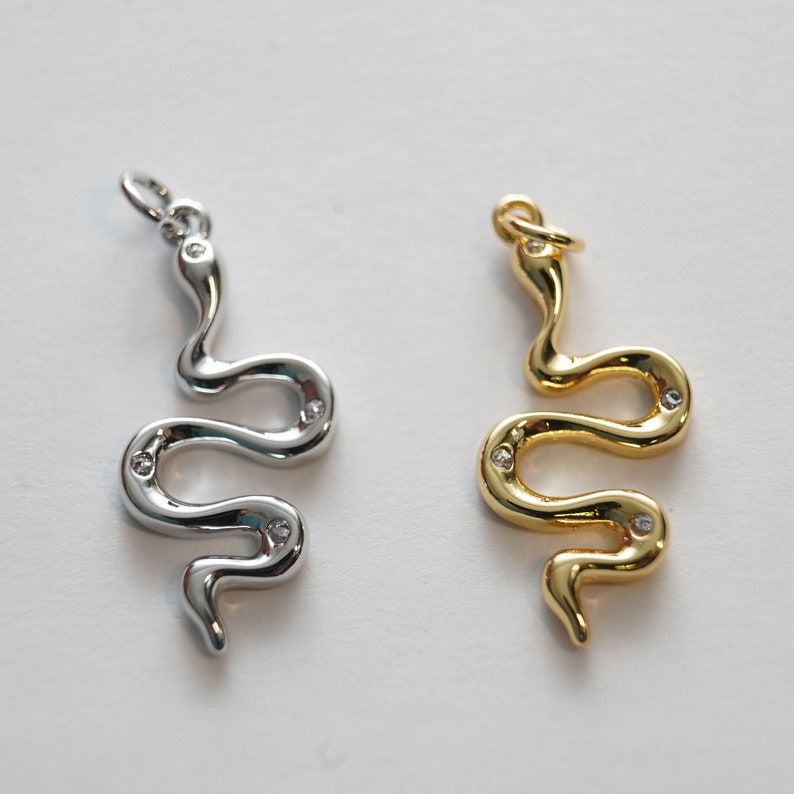 Op de afbeelding: Twee slangen-vormige hangers met heldere kristallen accenten. E&eacute;n hanger is zilver en de andere is goud.