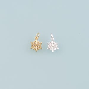 May include: Two snowflake charms, one gold-toned and one silver-toned, are displayed against a light blue background. The charms have intricate details and are likely made of metal. Each charm has a small loop for attaching to a necklace or bracelet.