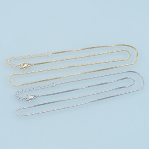 Puede incluir: Dos delicados collares de cadena, uno dorado y otro plateado, yacen uno al lado del otro sobre un fondo azul claro. Ambas cadenas tienen un cierre de caja.