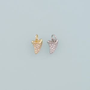 May include: Two pendant charms shaped like shark teeth. One is gold-colored and the other is silver-colored. Both charms are encrusted with small, clear crystals. The pendants have a loop at the top for attaching to a chain or bracelet.