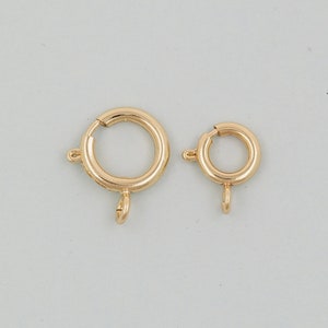 Op de afbeelding: Twee gouden metalen sprongringen met een veerclip. De grotere ring heeft een diameter van ongeveer 2,5 cm en de kleinere ring heeft een diameter van ongeveer 2 cm.