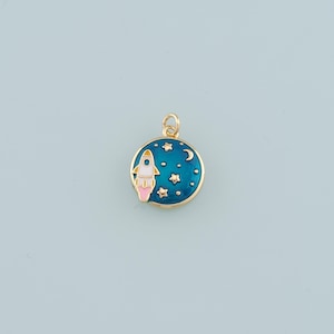 May include: A gold-colored round charm with a blue enamel background featuring a white and pink rocket ship, stars, and a crescent moon. The charm has a gold-colored loop at the top for attaching to a necklace or bracelet.