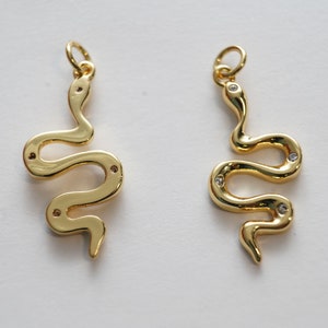Op de afbeelding: Twee gouden slangencharms met heldere kristallen accenten. De charms zijn in de vorm van een slang met zijn lichaam gekruld in een cirkel.