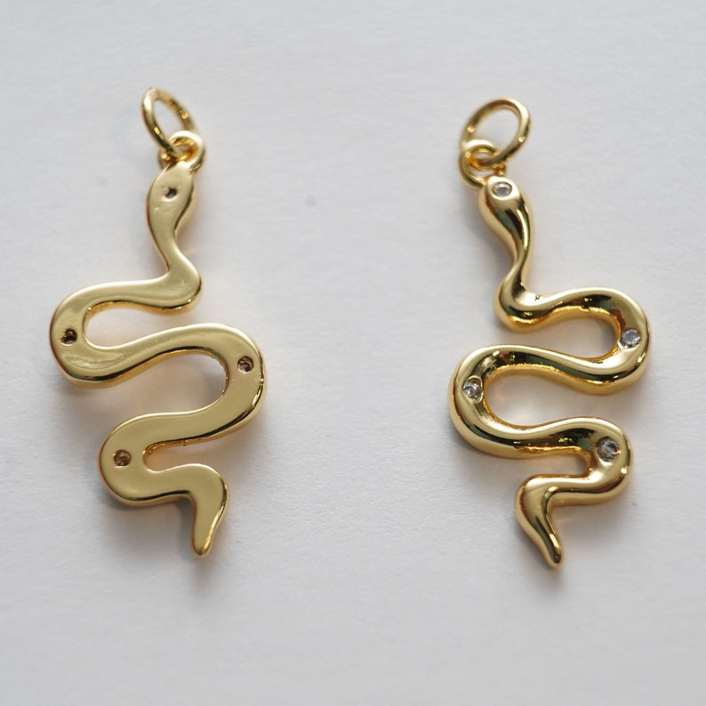 Op de afbeelding: Twee gouden slangencharms met heldere kristallen accenten. De charms zijn in de vorm van een slang met zijn lichaam gekruld in een cirkel.