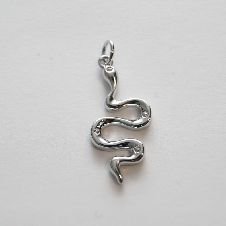 Op de afbeelding: Een zilveren slangenbedel met heldere kristallen accenten. De slang is opgerold en heeft een lus aan de bovenkant om aan een ketting of armband te bevestigen.