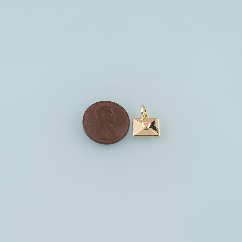 May include: A gold-colored charm in the shape of an envelope with a pink heart. The charm is next to a penny on a light blue background. The charm has a small loop for attaching to a necklace or bracelet.
