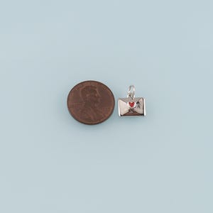 May include: A silver-colored charm in the shape of an envelope with a red heart. The charm is next to a penny for size comparison. The background is a light blue color.
