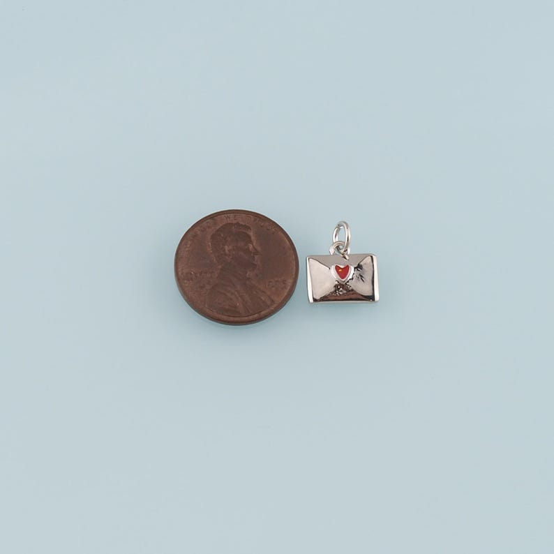 May include: A silver-colored charm in the shape of an envelope with a red heart. The charm is next to a penny for size comparison. The background is a light blue color.