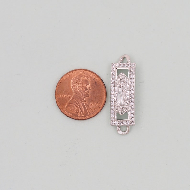 May include: Silver charm with a clear crystal border and a cutout image of the Virgin Mary inside. The charm is about the size of a UK penny.