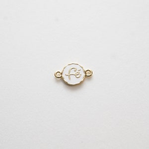 May include: A gold-toned charm with a white enamel circle featuring the word "Fé" in cursive script. The charm has two jump rings for attaching to jewelry.