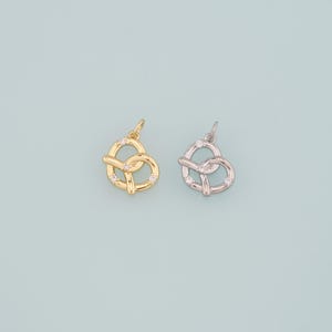 May include: Two pretzel-shaped pendants. One is gold-colored, the other is silver-colored. Both pendants have small, clear gemstones embedded in the design. The pendants are displayed against a light blue background.