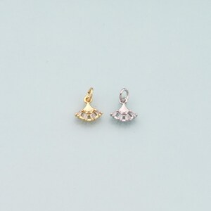 May include: Two small fan-shaped charms. One is gold-colored with clear, rectangular stones, and the other is silver-colored with similar stones. Both have a small loop at the top for attaching to jewelry. The background is a light blue.