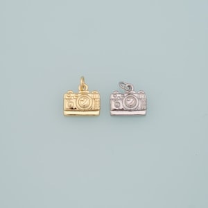 Peut inclure: Deux petits charms en forme d'appareil photo, l'un doré et l'autre argenté, sur un fond bleu clair. Les breloques présentent des détails de lentilles et de boutons, parfaits pour la création de bijoux ou l'artisanat.