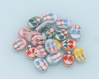 May include: A collection of oval-shaped ceramic beads with floral designs in shades of blue, red, green, and yellow. The beads are white with colourful painted details.