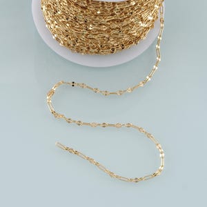 May include: A spool of gold-colored chain with a delicate, flat link design. The chain is partially unraveled, revealing its intricate pattern. The chain is likely used for jewelry making or other craft projects.