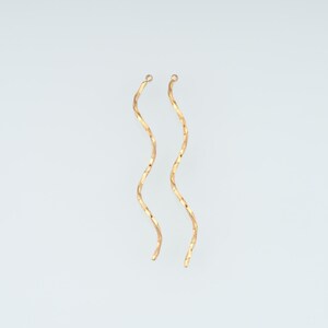 Può includere: Un paio di orecchini dorati lunghi e sottili caratterizzati da un design continuo e fluido a forma di onda o spirale. Ogni orecchino è composto da un singolo filo metallico sottile modellato in una curva a S allungata che pende verticalmente.