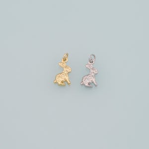 May include: Two small bunny charms, one gold-colored and one silver-colored, sit side-by-side against a light blue background. The charms are detailed with textured surfaces, and each has a small loop at the top for attaching to a chain or bracelet.