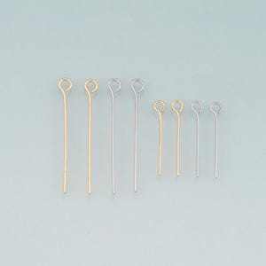 May include: An assortment of gold and silver-colored eye pins of varying lengths, arranged against a light blue background. The pins have a looped end and a straight wire shaft, ideal for jewelry making and crafting projects.
