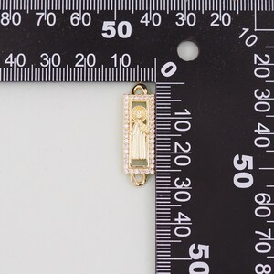 May include: Gold-toned charm with a clear crystal border, featuring a raised image of a saint inside. The charm is 3.8 cm tall and is measured against a ruler.