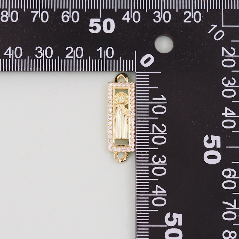 May include: Gold-toned charm with a clear crystal border, featuring a raised image of a saint inside. The charm is 3.8 cm tall and is measured against a ruler.
