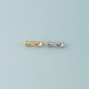 May include: Two small, decorative clasps. One is gold-toned, the other silver-toned. Both have a cylindrical shape with a central section adorned with small, clear gemstones. The clasps are designed for jewelry making or similar crafts.