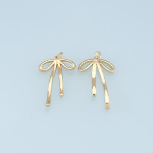 May include: Two gold-colored metal bow charms with a simple design. The charms are shaped like a bow with two long, thin tails.