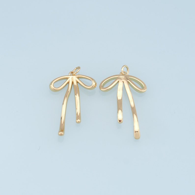 May include: Two gold-colored metal bow charms with a simple design. The charms are shaped like a bow with two long, thin tails.