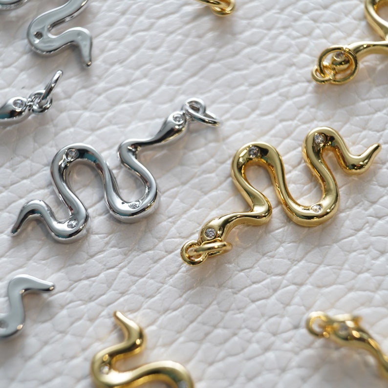 Op de afbeelding: Twee zilveren en twee gouden slangencharms met heldere kristallen accenten. De charms zijn op een witte achtergrond.