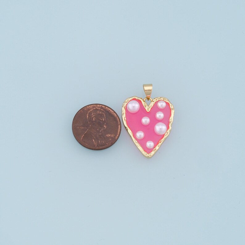 May include: A pink heart-shaped pendant with a gold border and white pearl accents. The pendant is about the size of a US penny.