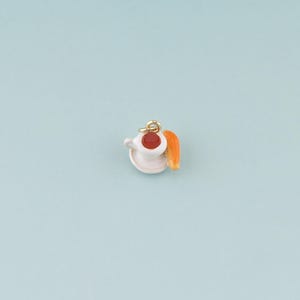May include: A small, white teacup charm with a saucer, filled with a red liquid, and a gold-colored loop for hanging. An orange-colored croissant is next to the cup. The charm is set against a light blue background.