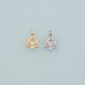 May include: Two Star of David pendants, one gold-toned and one silver-toned, each with a heart-shaped center. The hearts and the star points are embellished with small, clear gemstones. The pendants are displayed against a light blue background.