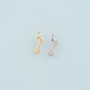 May include: Two dog bone charms, one gold-toned and one silver-toned, each encrusted with small, clear crystals. The charms are suspended from small loops, perfect for jewelry making or pet-themed crafts. The background is a light blue.