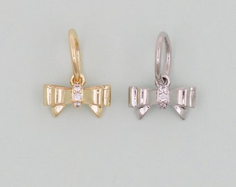 May include: Two bow-shaped charms, one gold-toned and one silver-toned, each with a small hoop and three clear accent stones. The charms are designed to be used as jewelry components, such as earrings or pendants.