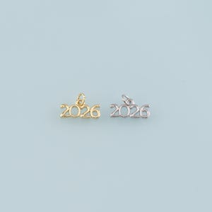 May include: Two small charms, one gold-colored and one silver-colored, each displaying the number "2026" in a delicate, cursive font. The charms have a small loop at the top for attaching to a necklace or bracelet. The background is a solid, light blue.