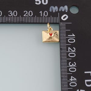 May include: A gold-colored envelope charm with a red heart detail. The charm is rectangular with a small loop at the top for attaching to a necklace or bracelet. The charm is approximately 0.5 inches in length.