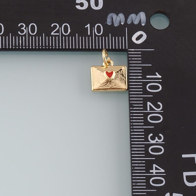 May include: A gold-colored envelope charm with a red heart detail. The charm is rectangular with a small loop at the top for attaching to a necklace or bracelet. The charm is approximately 0.5 inches in length.