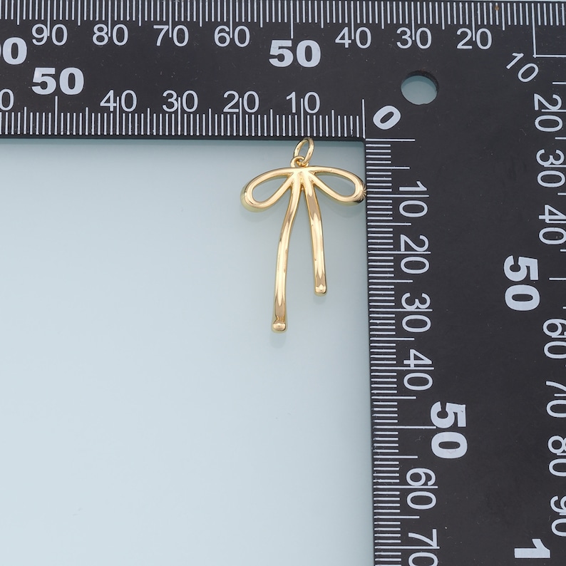 May include: A gold-colored bow charm pendant, measuring approximately 2.5 cm in length, with a simple design and a smooth finish.