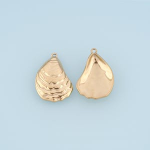 May include: Two gold-toned metal charms shaped like oyster shells. The left charm displays the textured, ridged exterior of a shell, while the right charm shows the smooth, concave interior. Each piece features a small circular loop at the top.