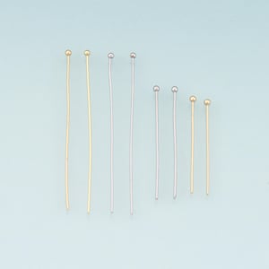 May include: Assortment of head pins in gold and silver tones. The pins have a small ball at the top and a straight wire shaft. The pins are arranged on a light blue background. The pins vary in length.