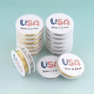May include: Several spools of jewelry wire in gold and silver tones are stacked and arranged on a light blue surface. The spools are labeled with "USA" and size measurements of 0.2mm, 0.3mm, and 0.6mm.