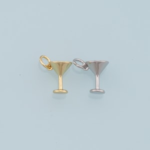 May include: Two small martini glass charms. One is gold-colored, the other is silver-colored. Both charms have a loop at the top for attaching to a necklace or bracelet. The charms are on a light blue background.