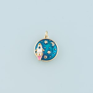 May include: Gold-toned charm with a blue enamel circle depicting a white rocket ship with pink flames, flying through a starry night sky with a crescent moon.