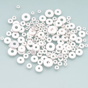 May include: A collection of small, silver-colored, flat, round beads with a hole in the center. The beads vary in size and are scattered across a light blue surface. These beads are suitable for jewelry making and crafts.